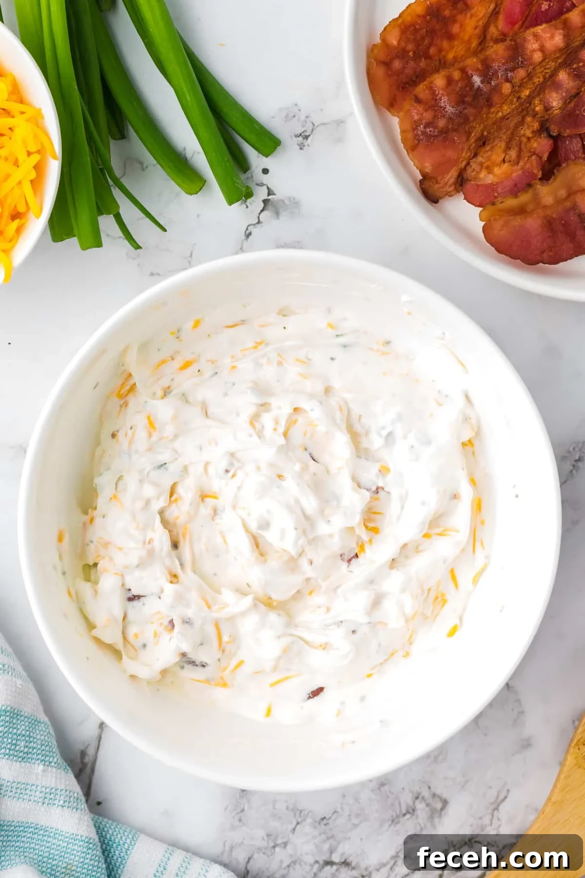 A white bowl filled with a sour cream based dip, with bowls of cooked bacon and cheese on the side.