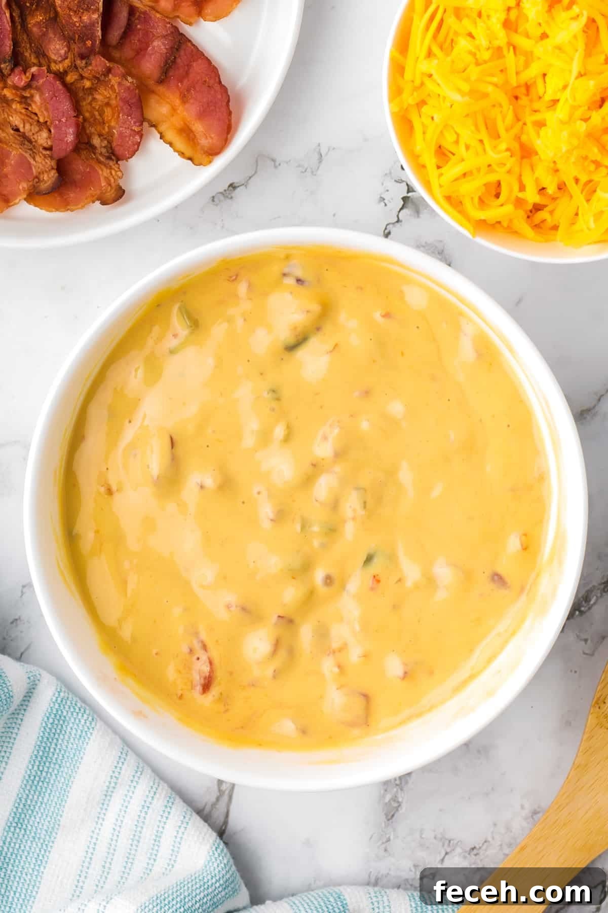 A large bowl of melted nacho cheese dip, with bowls of shredded cheese and bacon on the side.
