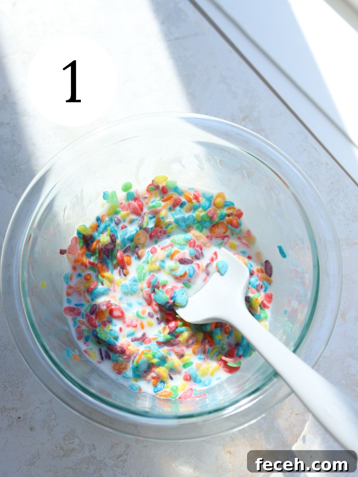 A glass bowl with fruity pebbles and milk and a white spatula.