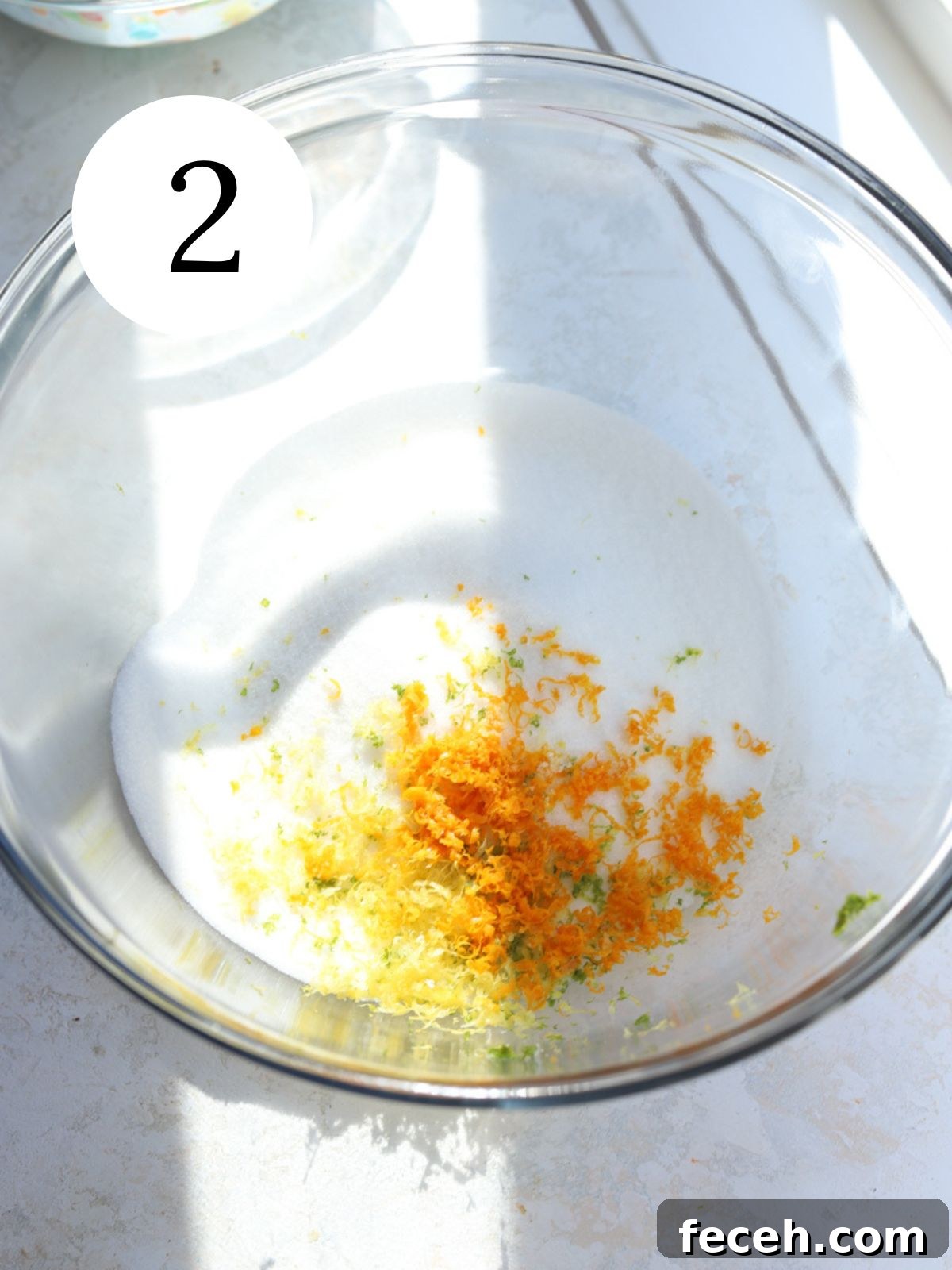 A glass mixing bowl with sugar, lemon zest, orange zest and lime zest.