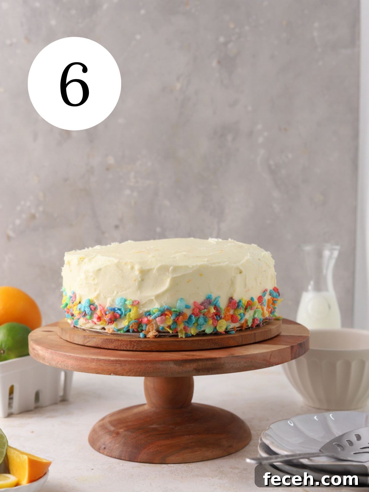 A fruity pebbles layer cake on a cake stand, a jug of milk, and some fruit.