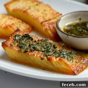 A close-up of a white plate with three golden-brown, crispy Domino Potatoes stacks, garnished with green herbs. A small bowl of herb-infused dipping sauce is beside them.
