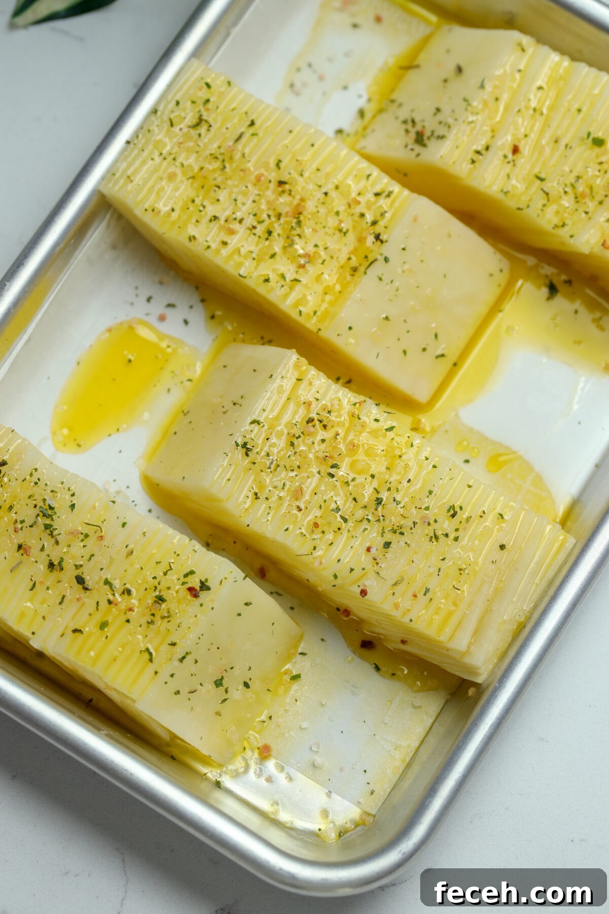 A metal baking tray filled with rectangular potato stacks, drizzled with olive oil and sprinkled with herbs. The sliced potato are arranged in rows and are ready to be baked.