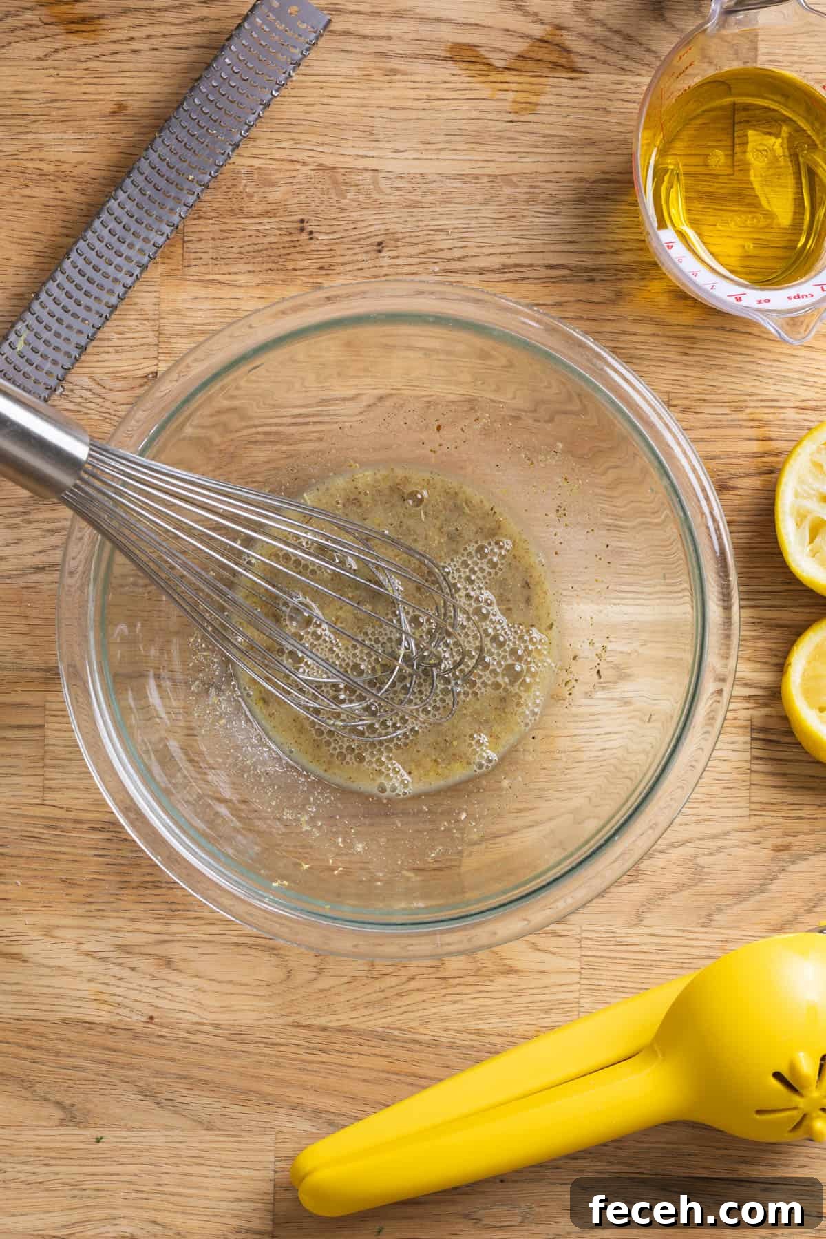 A whisk blending together ingredients for a lemon Dijon dressing in a glass bowl on a butcherblock countertop.