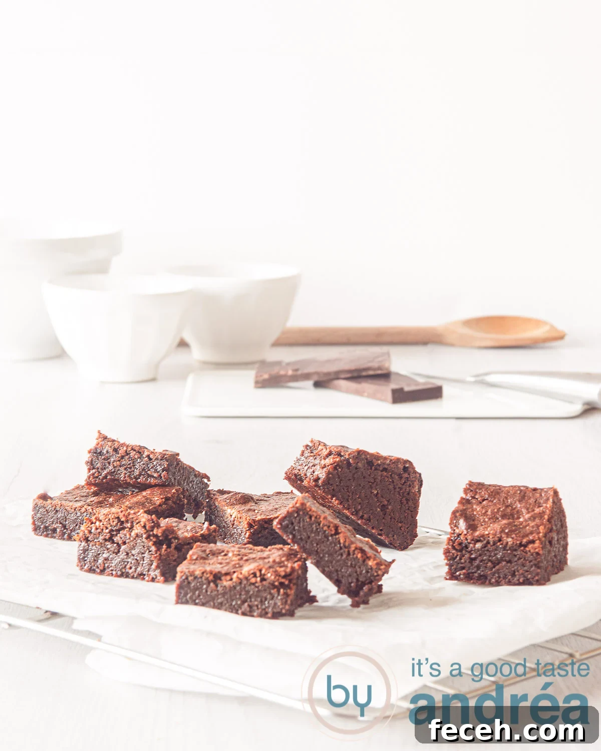 pieces of brownies scattered on a piece of baking paper. In the background some bowls, chocolate and wooden spoon