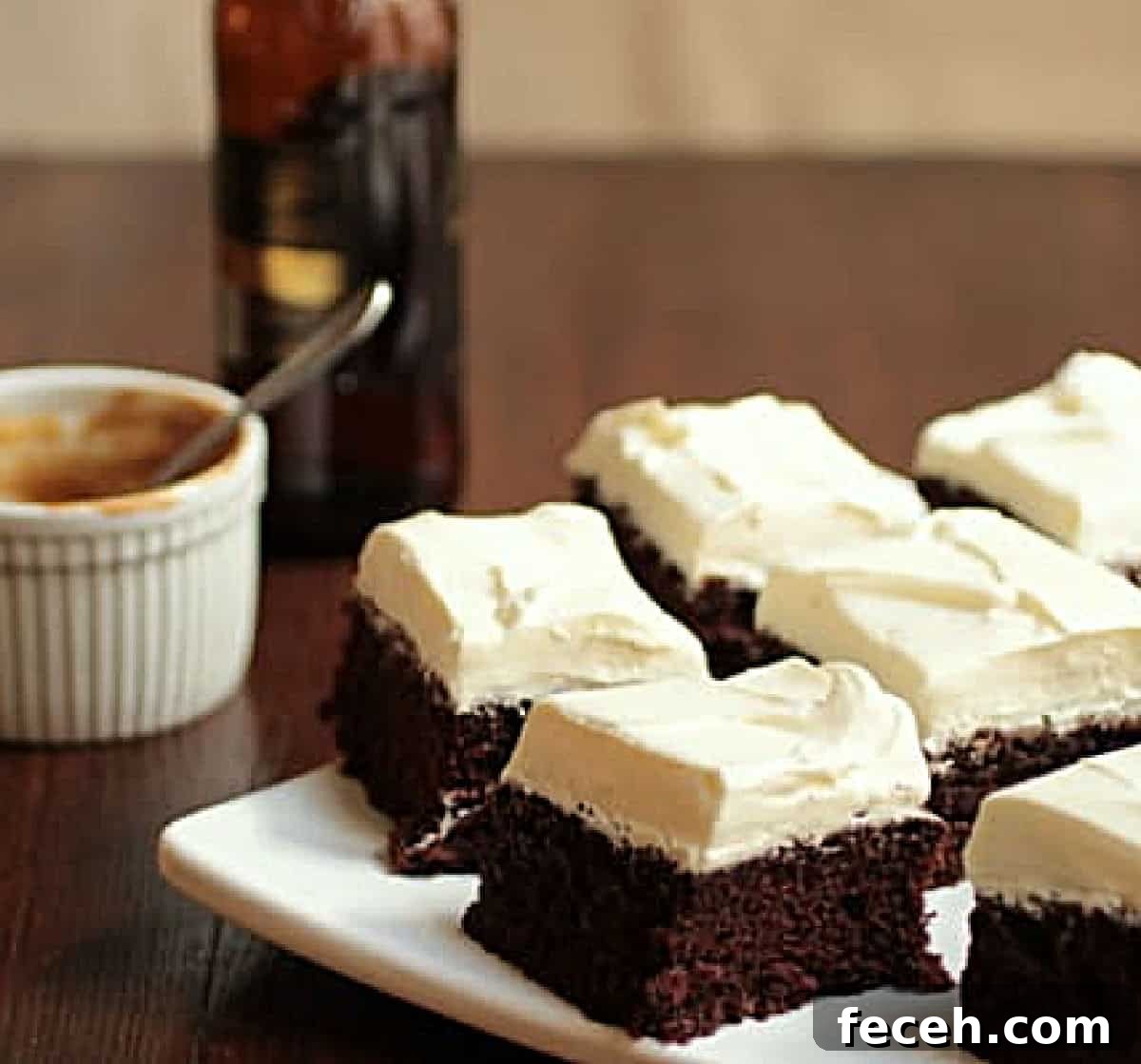 Cream topped chocolate cake squares on white plate, wooden table, stout bottle and white ramekin around
