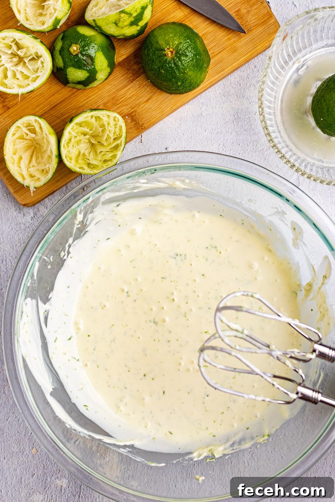 Key lime pie filling in a mixing bowl with mixing beaters hovering above it. Juiced, and zested limes are in the background on a cutting board.