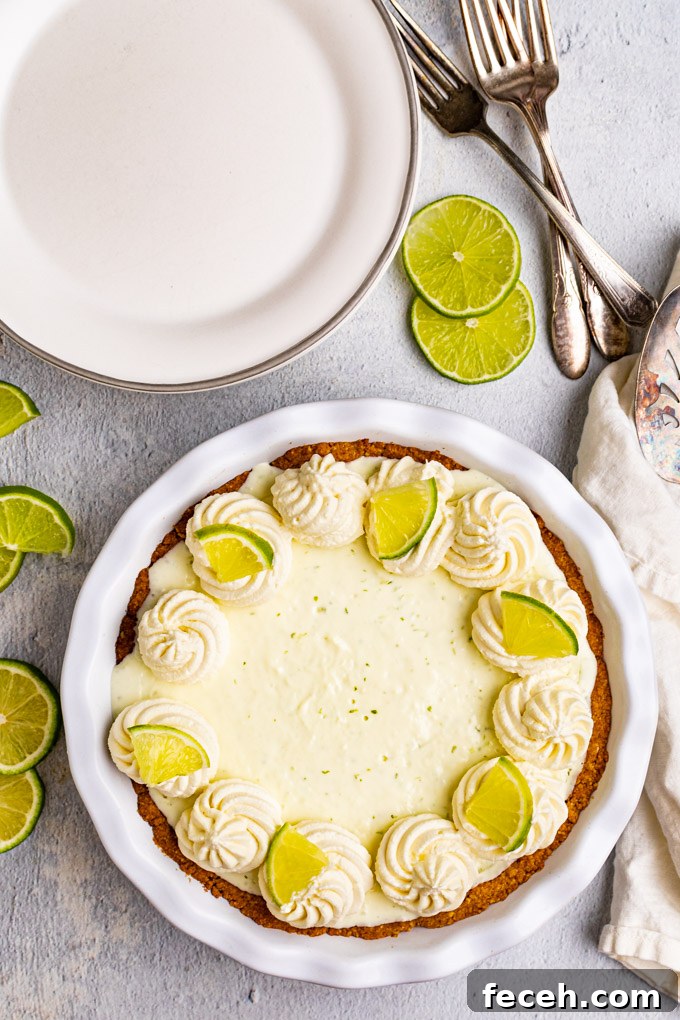 Overhead shot of the best key lime pie with rosettes of whipped cream decorating the outside, and lime slices on every other rosette. There are plain plates, a pie server, and forks in the background.