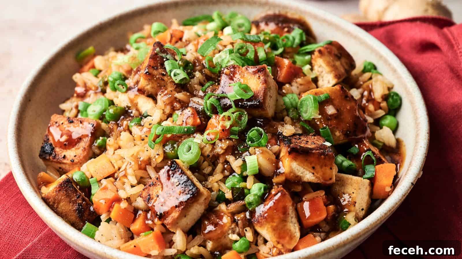 31 Asian Weeknight Dinners to Make Mealtime Exciting 26 A bowl of fried rice with grilled tofu, diced carrots, peas, chopped green onions, and a drizzle of dark sauce, served on a red cloth.