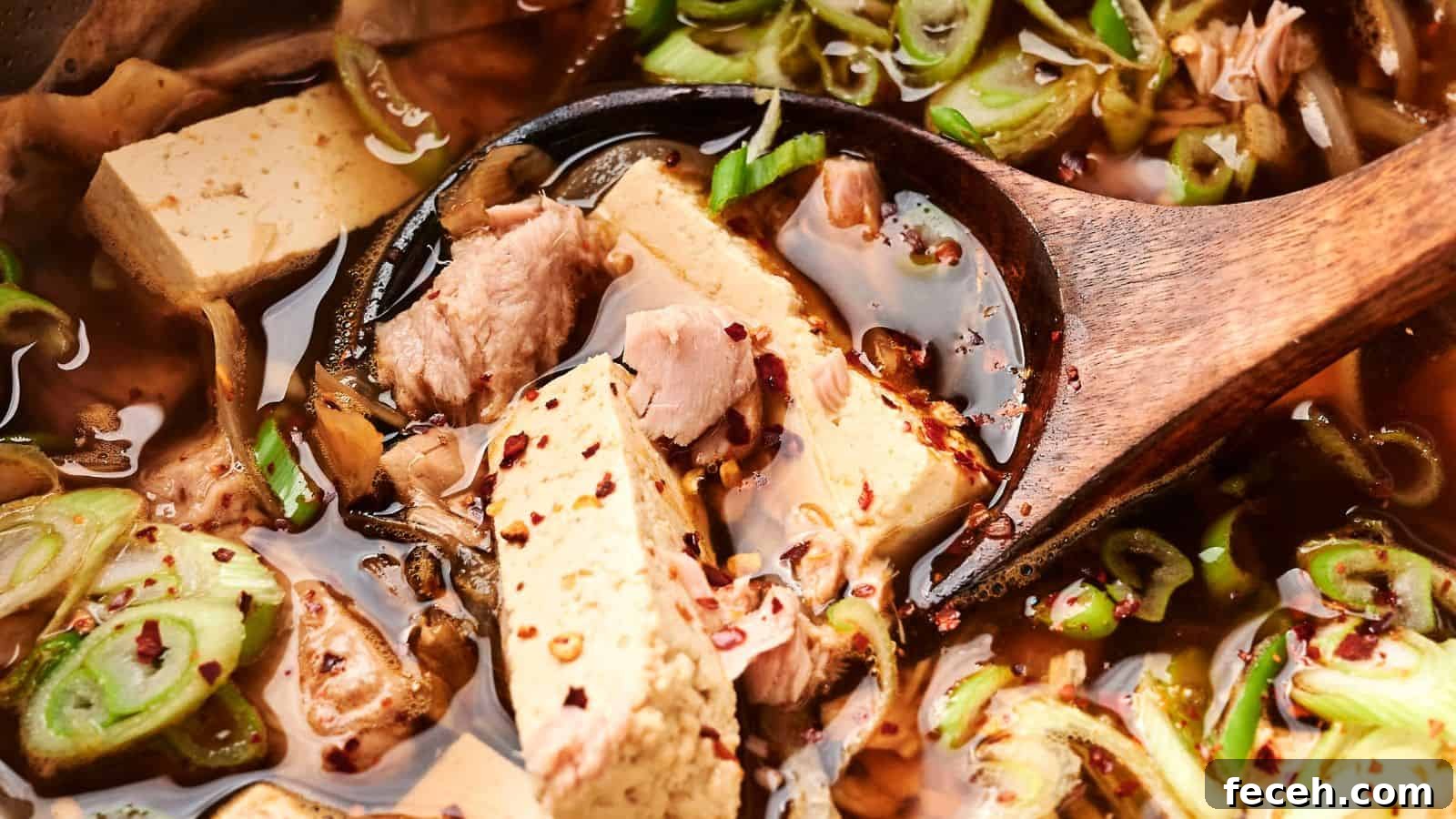 31 Asian Weeknight Dinners to Make Mealtime Exciting 28 Close-up of a wooden spoon in a vibrant kimchi stew with tofu and pork, garnished with sliced green onions and red pepper flakes.