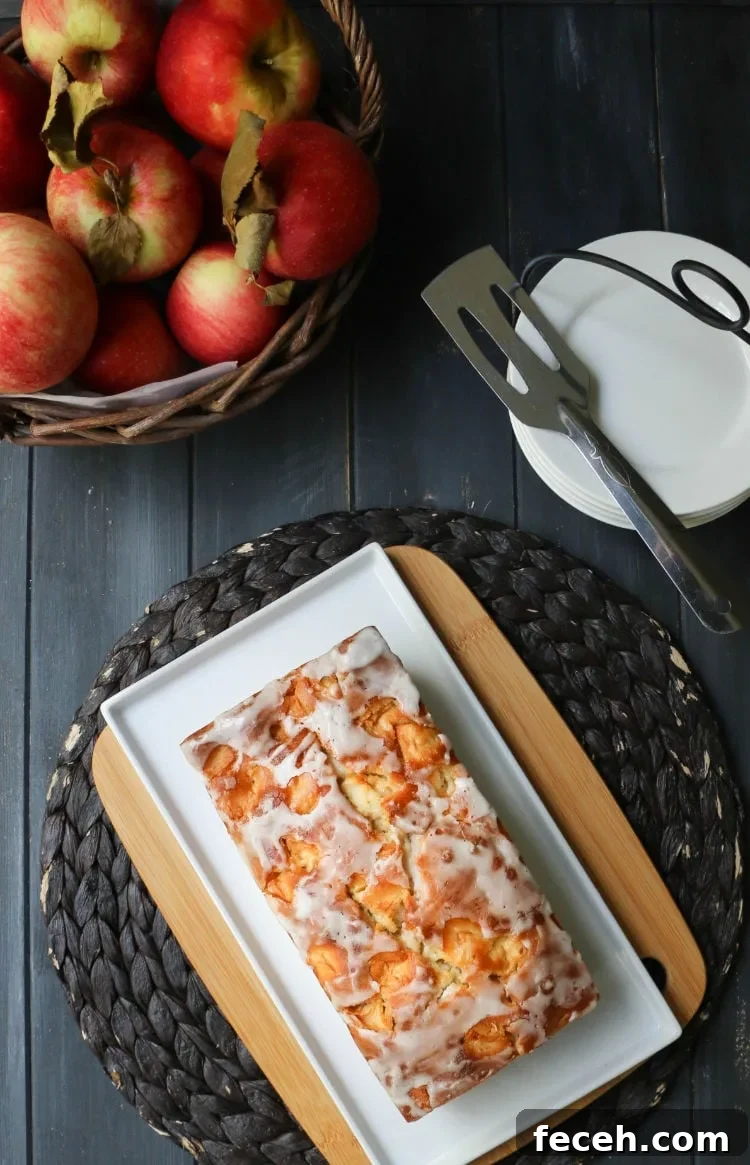 Top view of a whole loaf of Apple Fritter Bread on a white plate next to a basket of apples and a a stack of white plates with a serving spatula