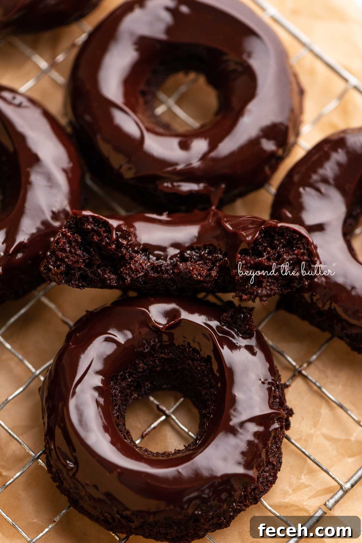 Homemade Baked Chocolate Donuts Recipe — Soft & Fudgy Treats 3 Half of a homemade baked chocolate donut topped with chocolate ganache on a wire cooling rack with more donuts | © Beyond the Butter®