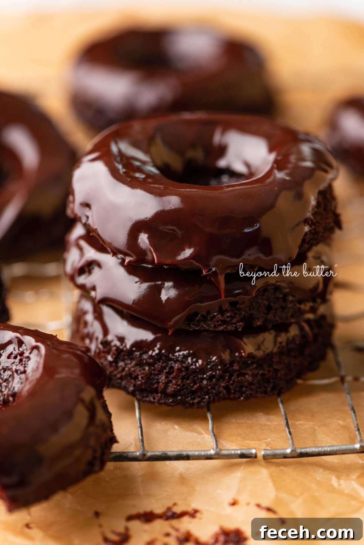 Homemade Baked Chocolate Donuts Recipe — Soft & Fudgy Treats 5 Stacked homemade baked chocolate donuts topped with chocolate ganache on a wire cooling rack | © Beyond the Butter®