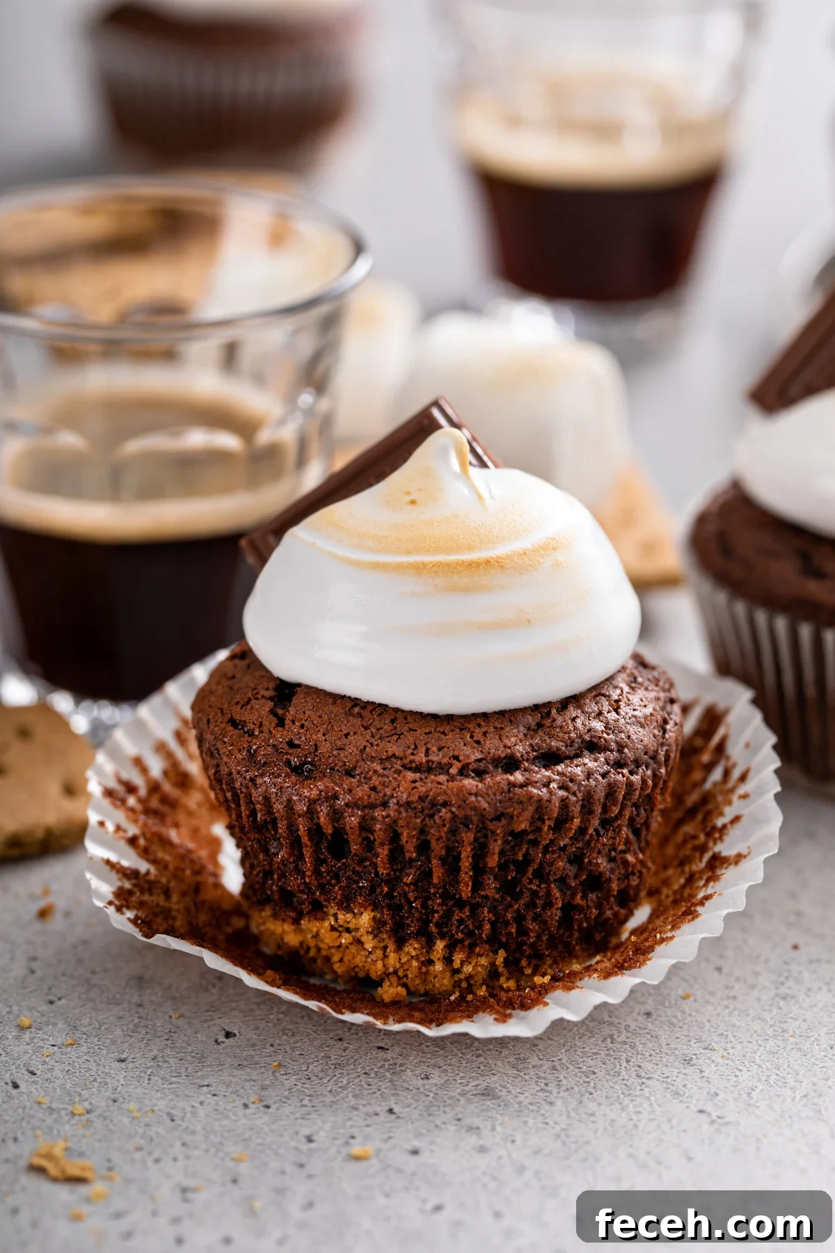 S'mores Cupcakes Recipe: Gooey Campfire Chocolate & Toasted Marshmallow 2 Unwrapped s'mores cupcake set on a gray countertop. A cup of espresso and more cupcakes are visible in the background.