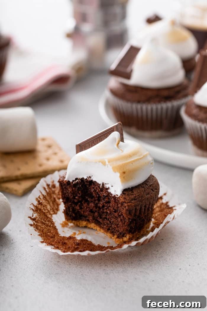 S'mores Cupcakes Recipe: Gooey Campfire Chocolate & Toasted Marshmallow 10 Unwrapped s'mores cupcake with a bite taken out of it. A platter of more cupcakes is visible in the background.