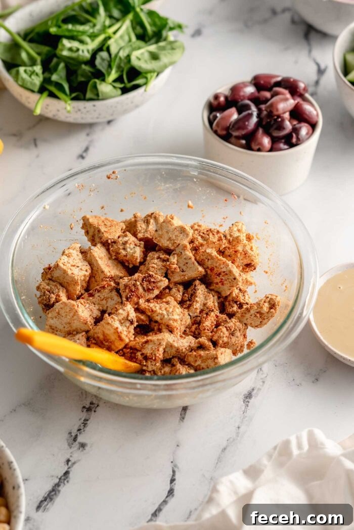 Firm tofu torn into pieces in a bowl and toss with a variety of spices. A small spatula rests in the bowl and there is a bowl of olives and a bowl of spinach in the background.
