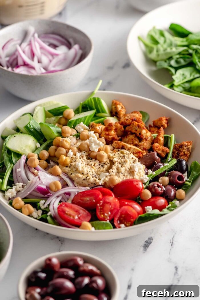 Healthy looking bowl with spinach, tofu, hummus, red onion, tomato, olives, cucumber and chickpeas. There are smaller bowls of sliced red onion, spinach and olives beside the bowl.