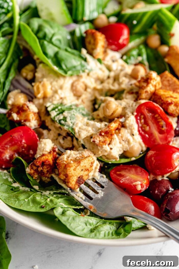 Fork resting in a bowl with spinach, tomatoes, olives, crispy tofu bits, hummus and chickpeas.