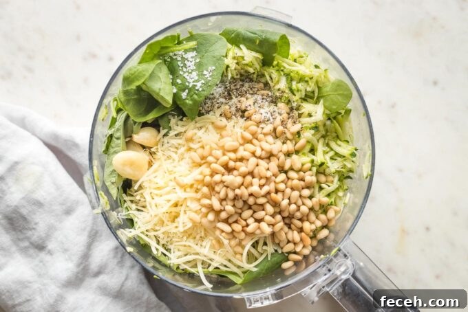 Grated zucchini, spinach and basil, and seasonings in the bowl of a food processor, not yet blended.