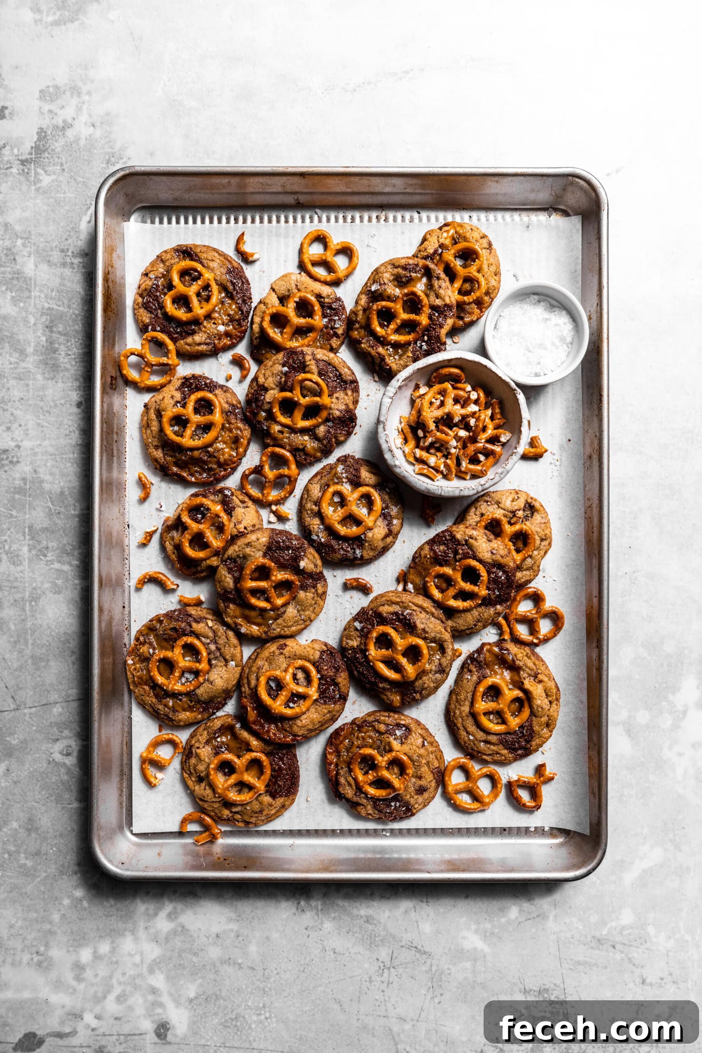 Freshly baked Salted Caramel Pretzel Chocolate Chip Cookies on a baking sheet next to small bowls of chopped pretzels and flaky sea salt