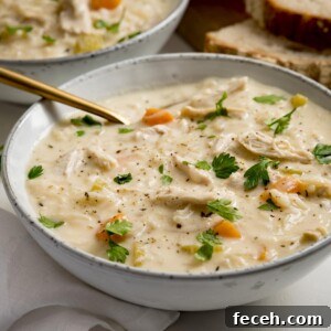 Square image of a bowl of chicken and rice soup on a light background. There is a gold spoon in the bowl. There is a further bowl and some bread slices in the background.