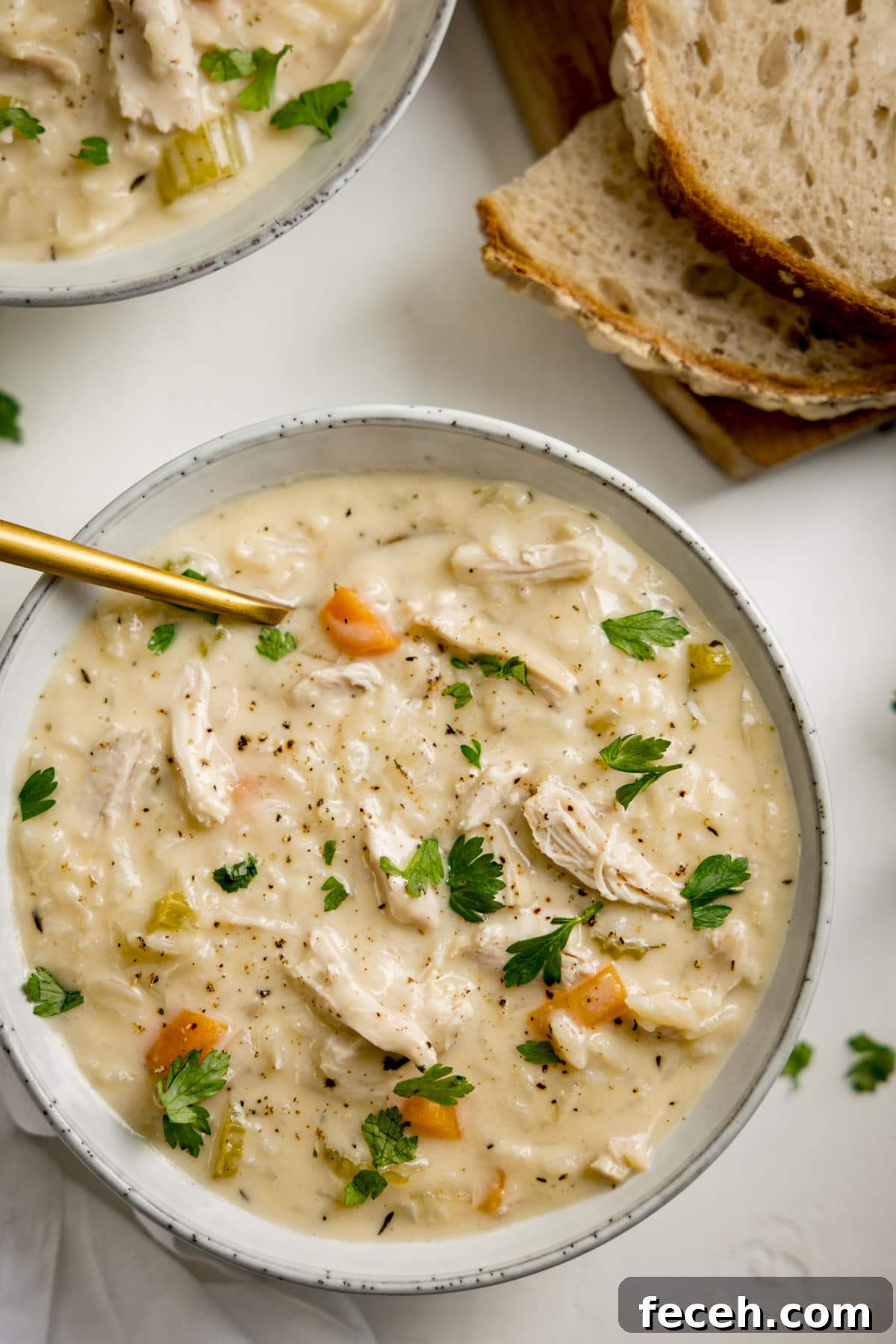 Overhead image of a light grey bowl filled with chicken and rice soup on a light surface. There is a gold spoon in the soup. There is a further bowl of soup and some slices of bread at the top of the frame.