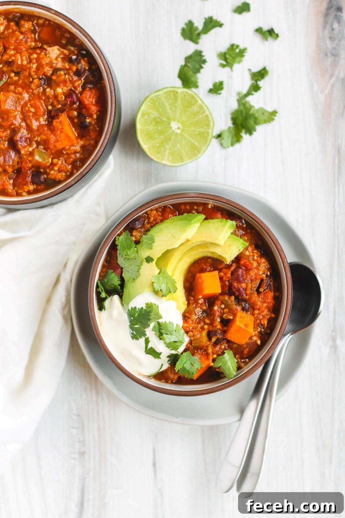 Sweet potato and quinoa chili in a bowl topped with avocado slices, cilantro, and sour cream with 2 spoons on the side and a separate bowl of chili in the back.