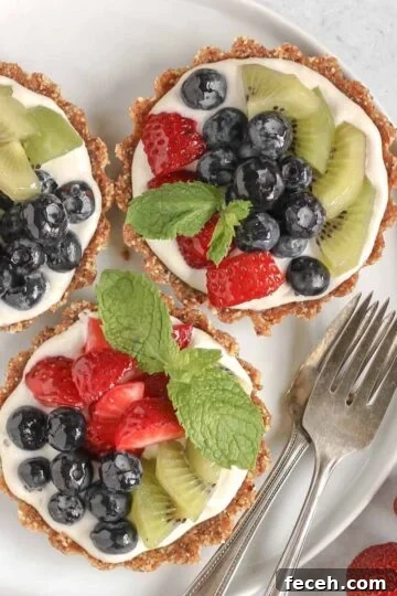 Three mini fruit tarts on a small white plate with 2 vintage forks on the plate and fresh berries off to the side.