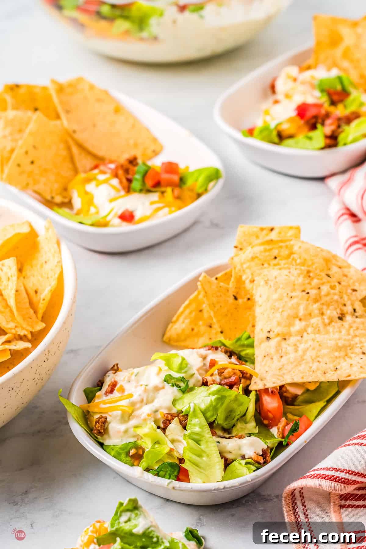 BLT Dip in a rectangle dish with chips on the side, on a table with other plates of the same thing.