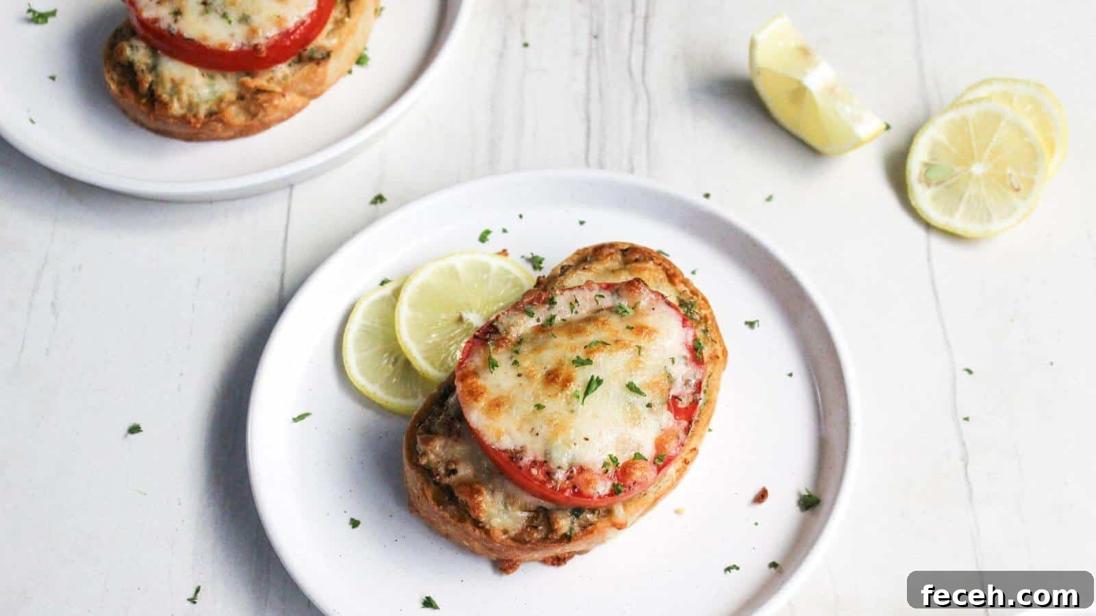Two plates of crab melt with sliced tomatoes and melted cheese, garnished with lemon slices on a white wooden surface.