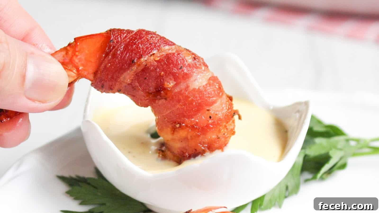 A hand holding a bacon-wrapped shrimp dipped in a small bowl of sauce, with a green leaf garnish on the side.