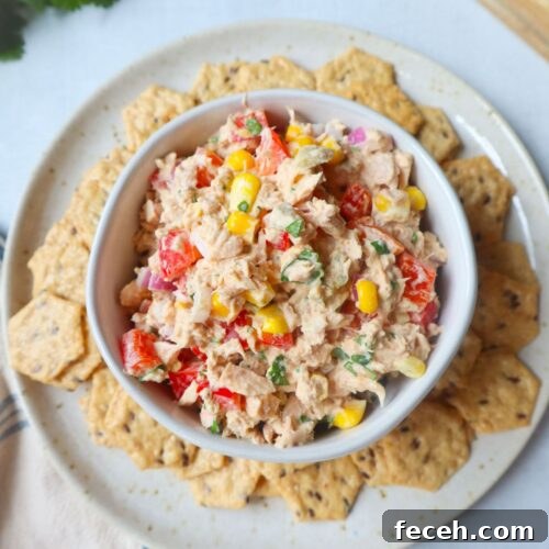 white bowl surrounded by crackers with tuna corn salad topped with herbs, red bell pepper, and onion