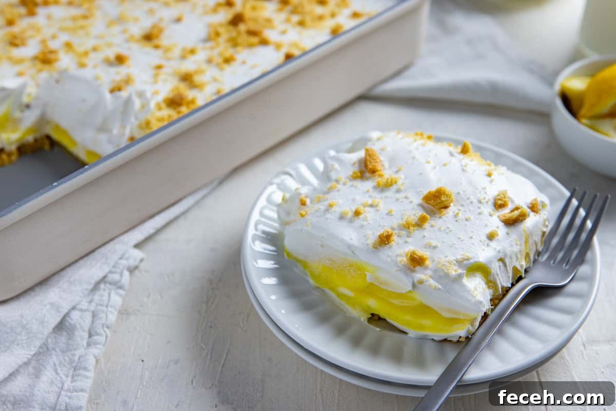 Creamy Lemon Lush Dessert Recipe for Every Occasion 12 A serving of lemon lush on a white plate with a fork on the side. The pan of lemon lush is in the background along with a bowl of sliced lemon wedges.