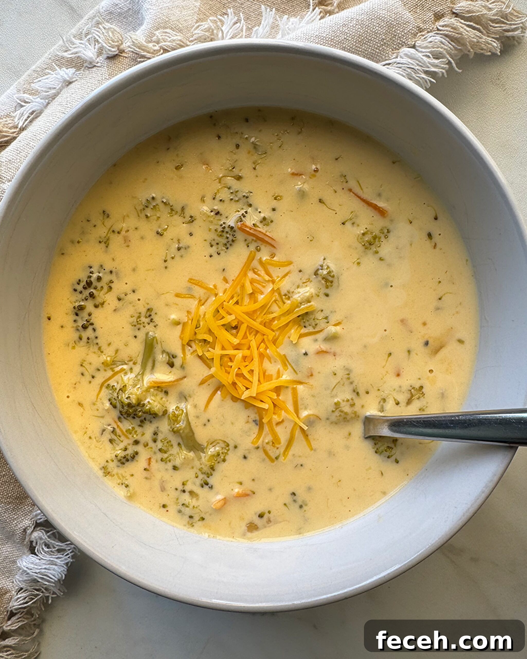 overhead shot of Broccoli Cheddar Soup in a white bowl garnished with cheddar cheese with a silver spoon