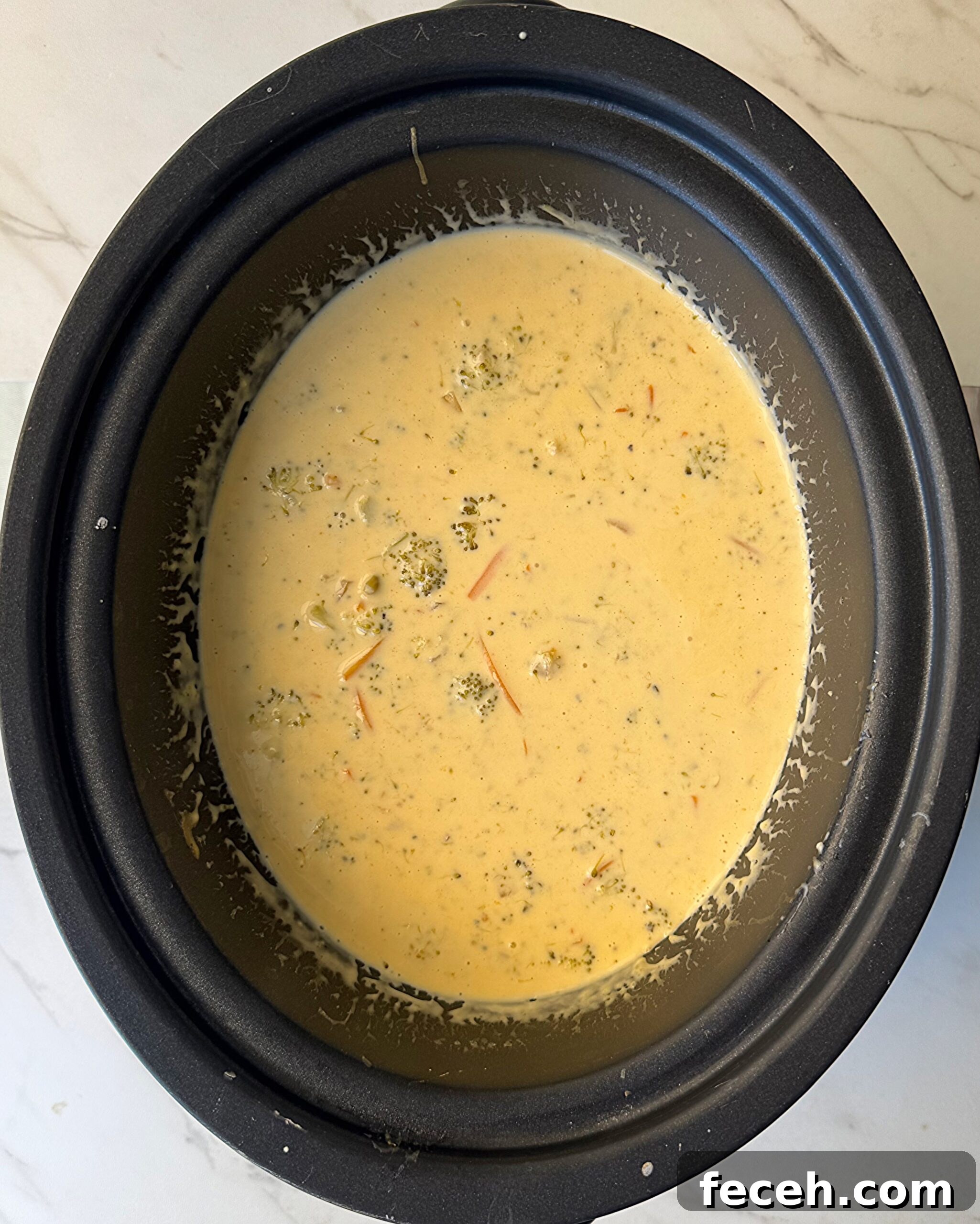 overhead shot of cooked Broccoli Cheddar Soup in a black slow cooker with cream and cheese added