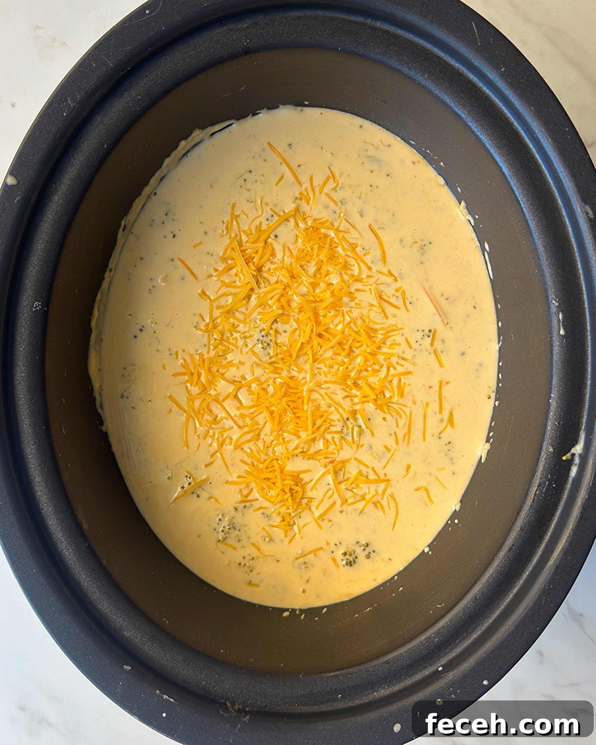 overhead shot of cooked Broccoli Cheddar Soup in a black slow cooker with garnished with shredded cheddar cheese