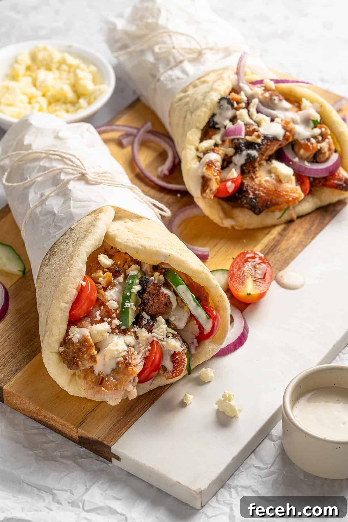 Two grilled pita wraps filled with chicken, tomatoes, cucumbers, red onions, and feta cheese drizzled with a creamy sauce, placed on a wooden board. A bowl of crumbled feta and a small dish of sauce are next to the wraps.