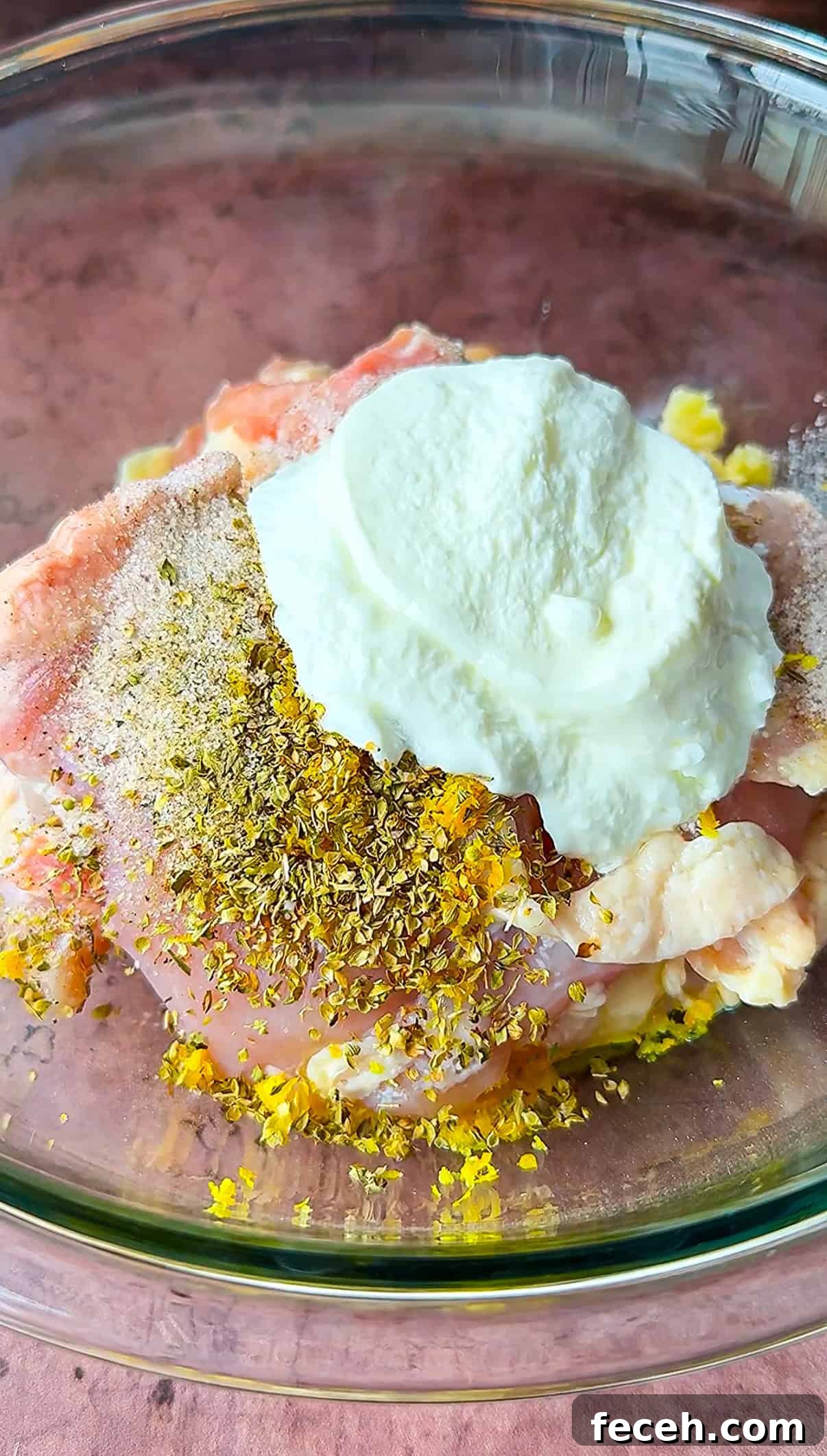 Raw chicken in a bowl is topped with ground spices, fresh lemon zest, and a dollop of plain yogurt. The background is a reddish-brown textured surface.