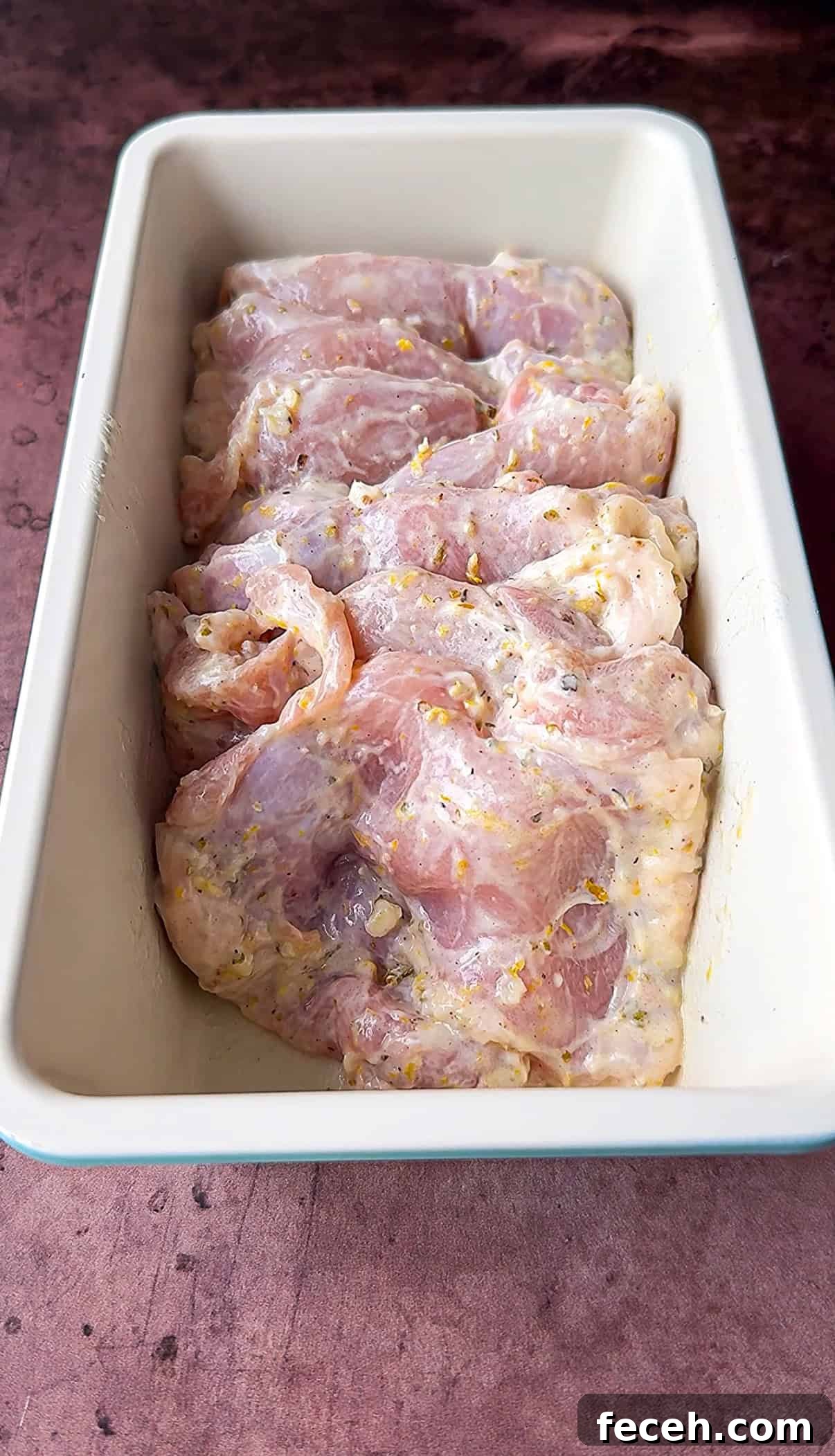 Raw chicken marinating in a rectangular pan, covered with a mixture of herbs and spices. The marinade appears to contain visible pieces of garlic and other seasonings. The pan is placed on a dark, textured surface.