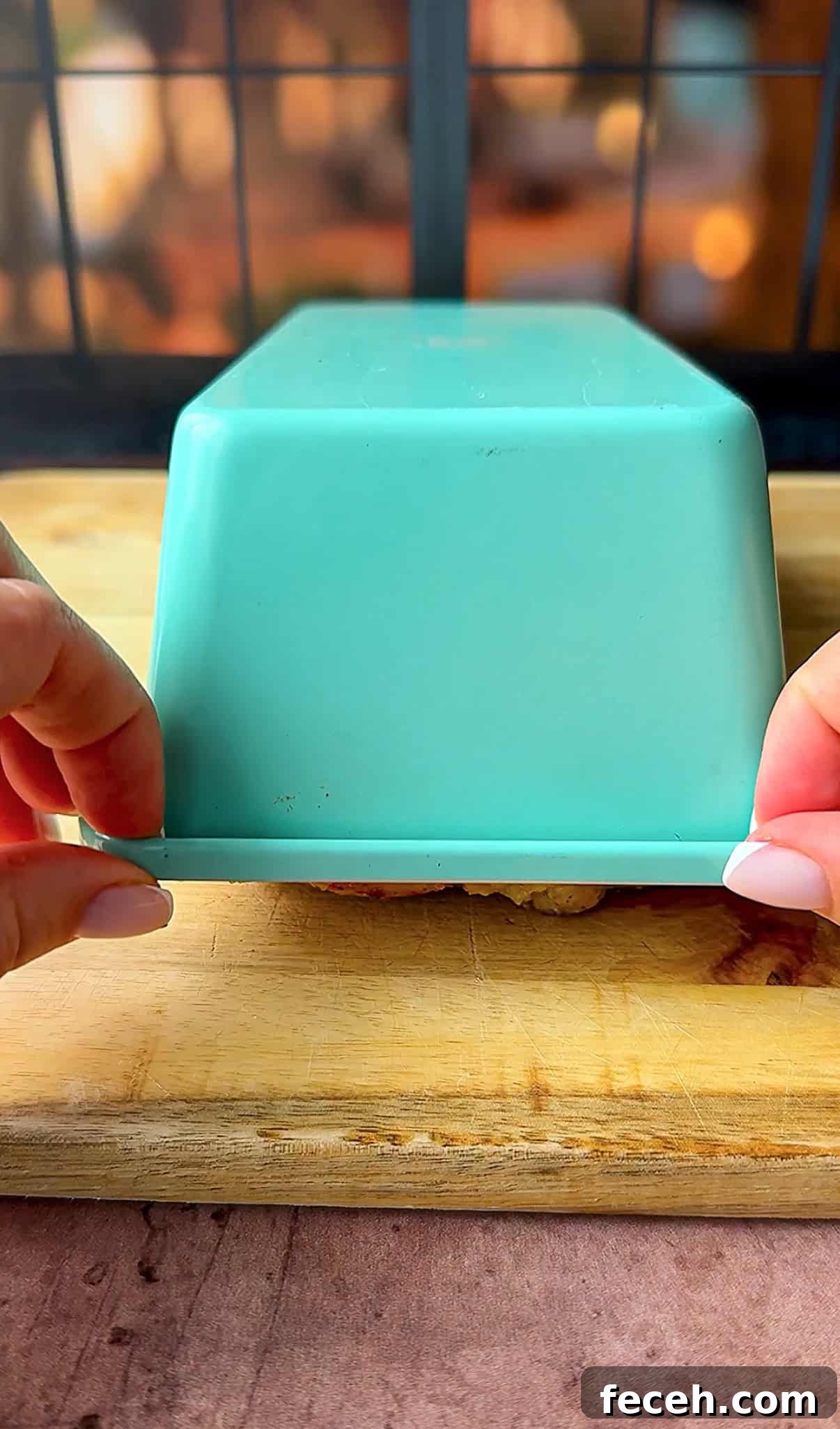 Two hands lift the edge of an upside-down, teal rectangular container placed on a wooden cutting board. The background features a black window frame with blurred outdoor scenery.