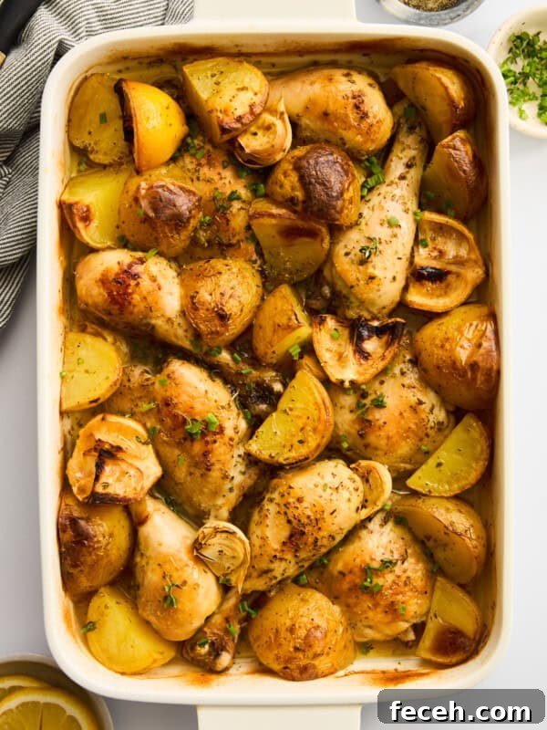 A white baking dish filled with roasted chicken drumsticks, potato wedges, and garlic cloves, garnished with herbs. The dish sits on a white surface with a lemon slice, chopped herbs, and a bowl of pepper nearby.