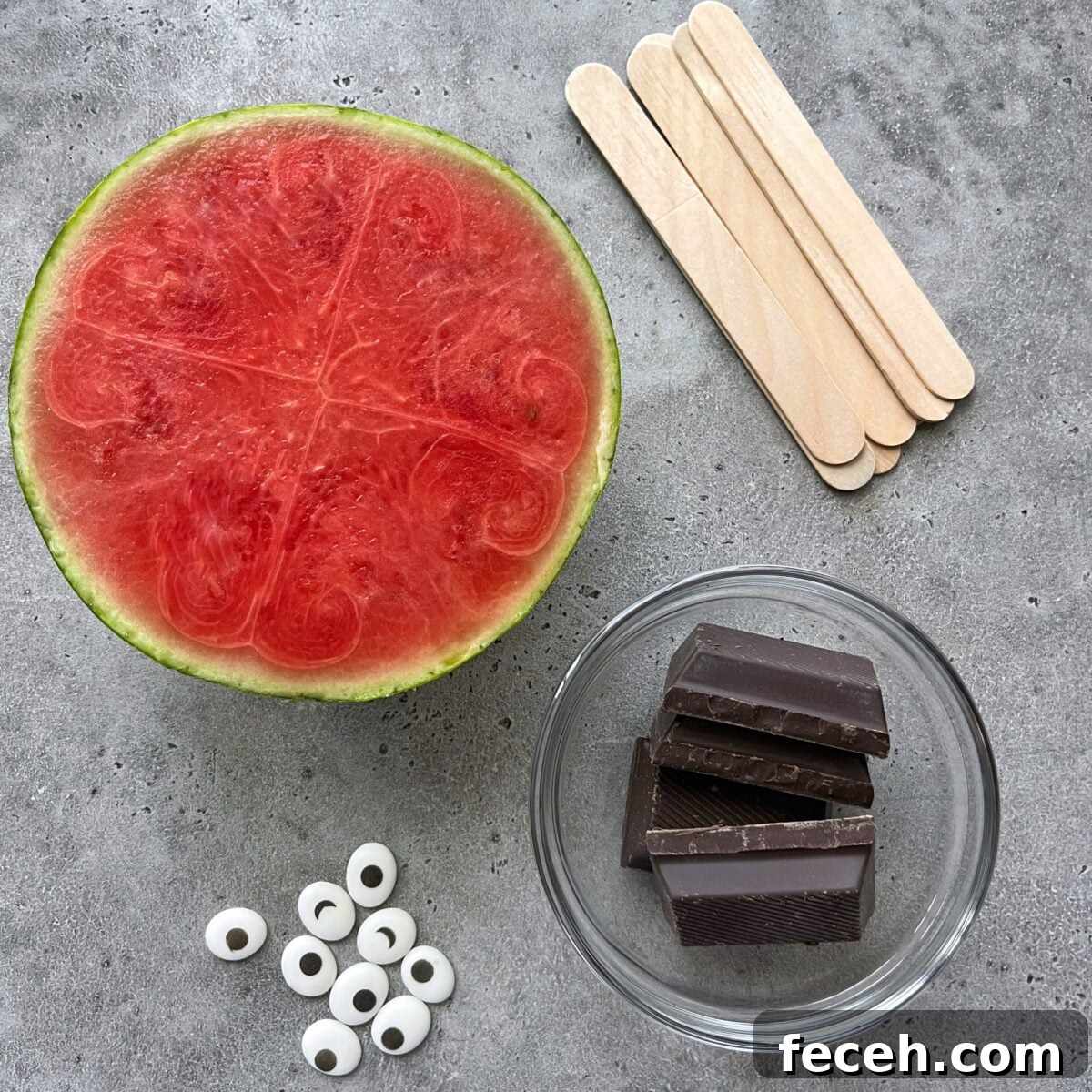 Half a watermelon, wooden popsicle sticks, a glass bowl with chocolate pieces, and candy eyes arranged on a gray surface.