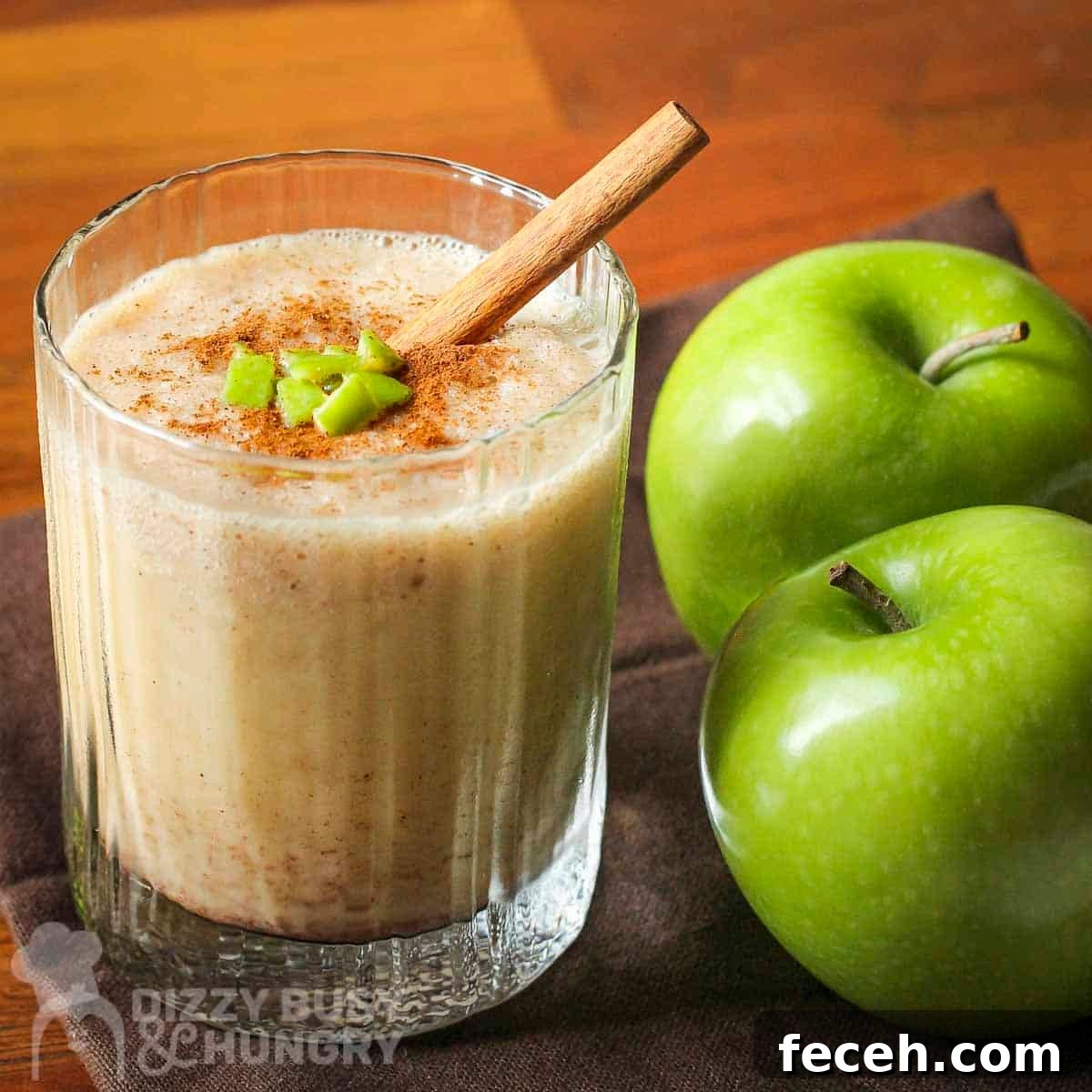 Side shot of a clear glass with apple smoothie garnished with cinnamon, small apple chunks, and a cinnamon stick with two granny smith apples on the side.