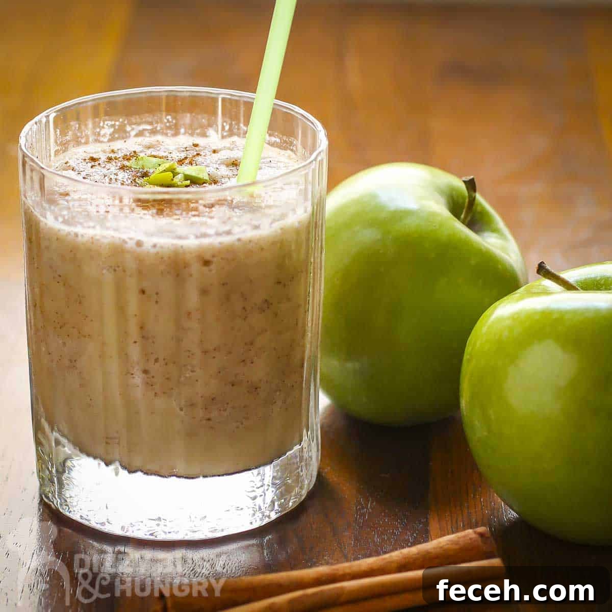 Side shot of a clear glass with apple smoothie garnished with cinnamon, small apple chunks, and a cinnamon stick with two granny smith apples on the side.