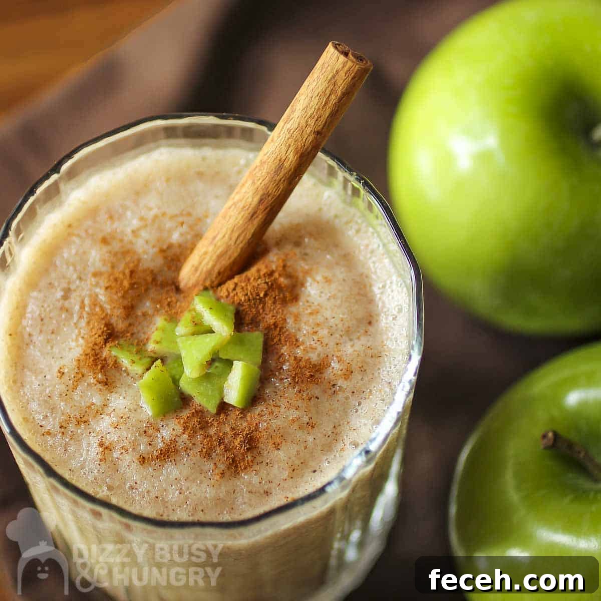 Overhead close up shot of a clear glass with apple smoothie garnished with cinnamon, small apple chunks, and a cinnamon stick with two granny smith apples on the side.