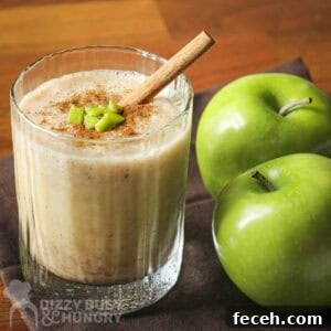 Side shot of a clear glass with apple smoothie garnished with cinnamon, small apple chunks, and a cinnamon stick with two granny smith apples on the side.