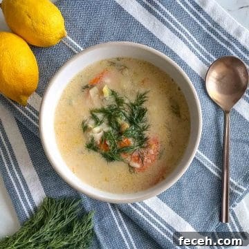 A bowl of Greek Chickpea Soup garnished with dill sits on a blue and white linen with a spoon, fresh dill, and lemons.