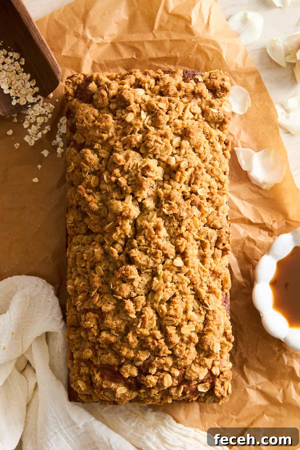 Apple Banana Bread with Crunchy Oat Crumble Topping 3 A loaf of bread or cake with a golden oat crumb topping sits on brown parchment paper, surrounded by oats, a white cloth, flower petals, and a small bowl of syrup.
