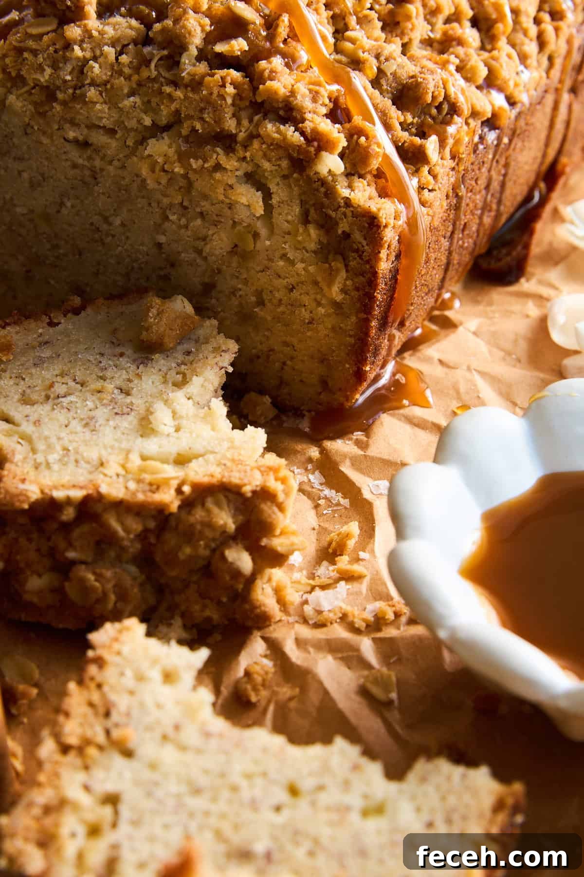Apple Banana Bread with Crunchy Oat Crumble Topping 4 Close-up of sliced crumb cake with a streusel topping and caramel drizzle, resting on brown parchment paper. A white dish with caramel sauce sits nearby. Crumbs are scattered around the slices.
