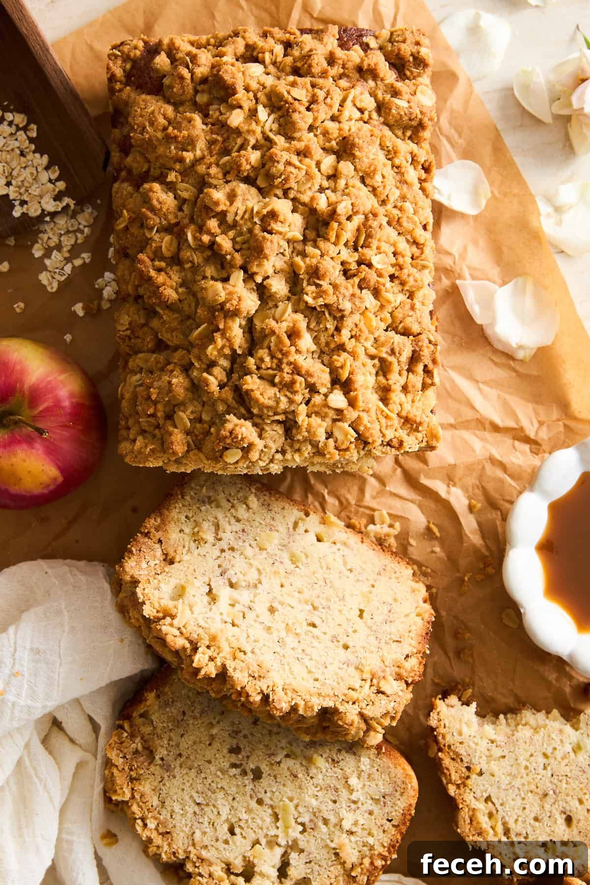 Apple Banana Bread with Crunchy Oat Crumble Topping 5 A loaf of apple crumble bread on parchment paper, partially sliced with three pieces cut. An apple, a white cloth, a bowl of caramel sauce, oats, and flower petals are placed around the bread.